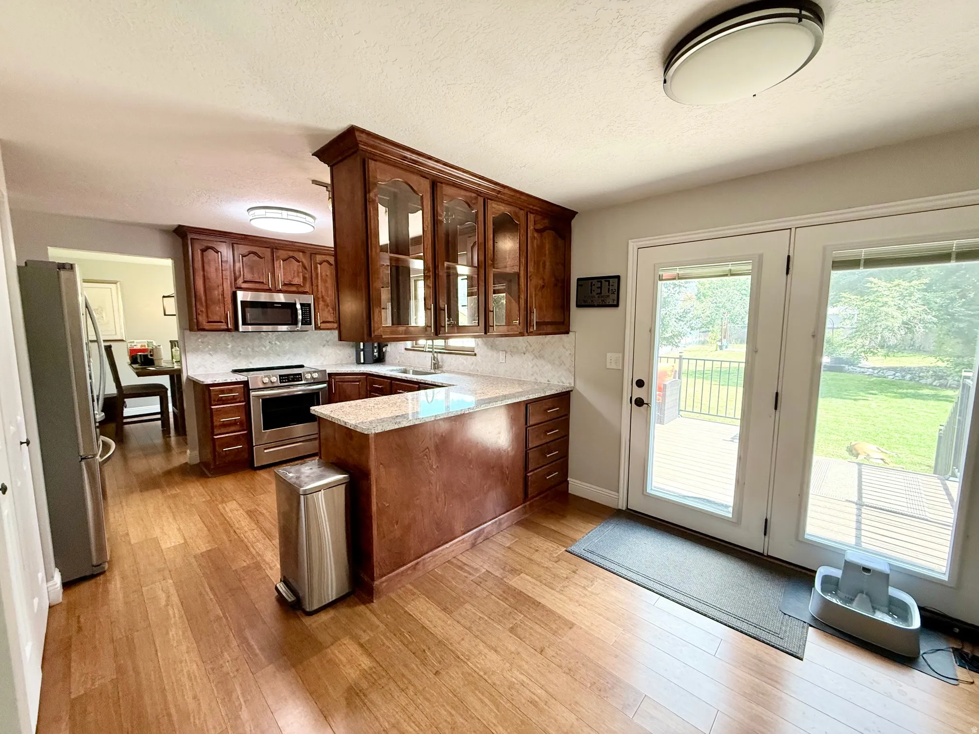 Kitchen with wood-finish flooring, wood cabinetry with glass accents, light-toned countertops, stainless steel appliances, and a full-light patio door leading to an outdoor deck