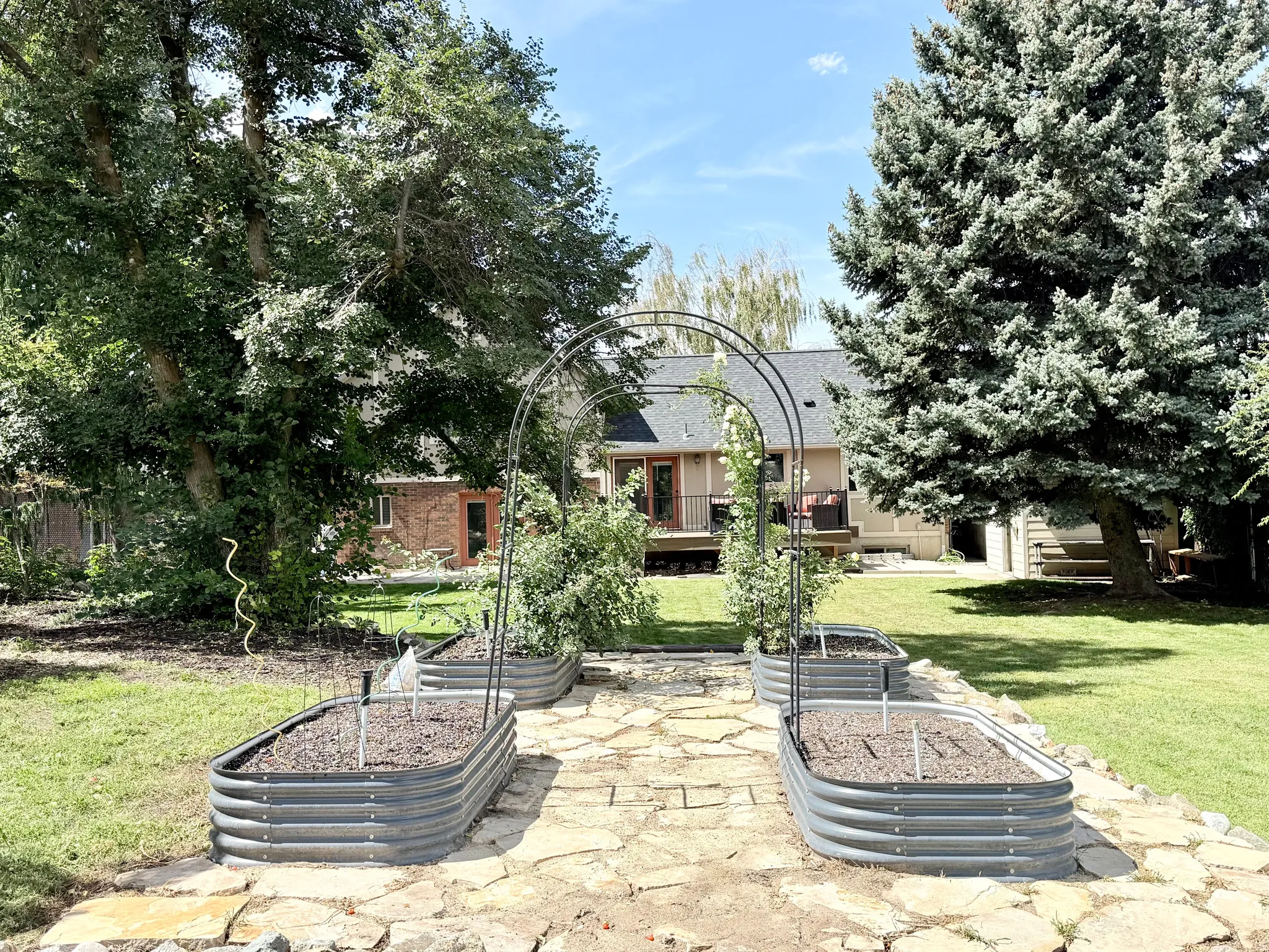 Expansive backyard featuring a stone pathway, multiple raised garden beds, mature trees, and a ground-level patio