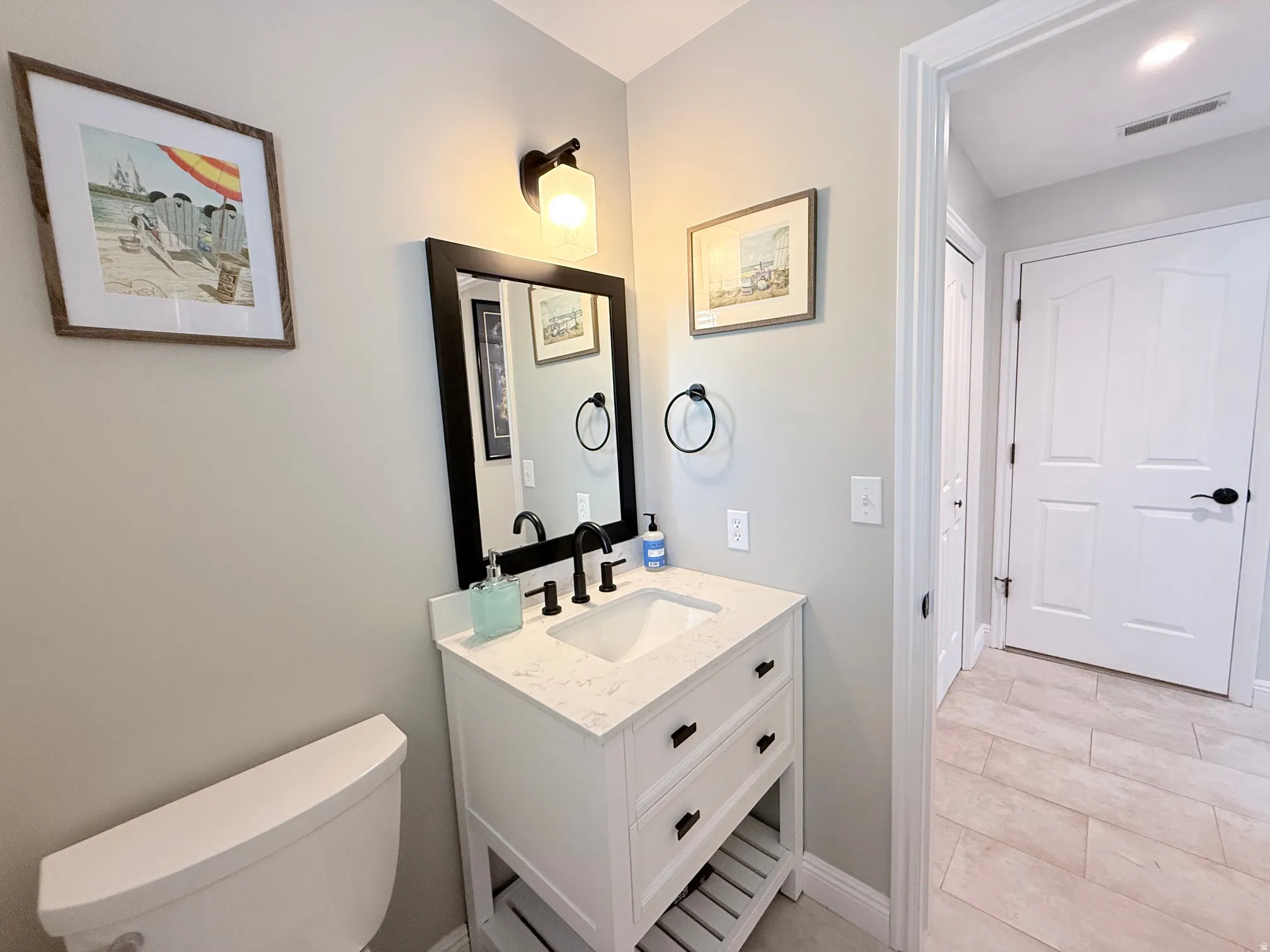 Vanity with a white countertop and integrated sink, black-framed mirror, contemporary wall sconce, and wood-finish tile flooring