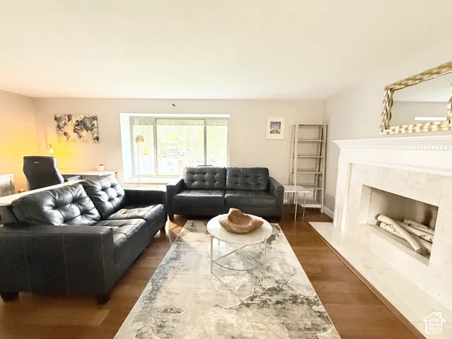Living area featuring wood-finish flooring, a fireplace with a light-toned stone surround, a prominent mantle, and a large picture window