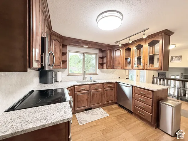 Kitchen featuring stained wood cabinetry, light-toned stone countertops, stainless steel appliances, glass-front upper cabinet doors, and wood-finish flooring