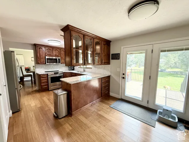 Kitchen with wood-finish flooring, wood cabinetry with glass accents, light-toned countertops, stainless steel appliances, and a full-light patio door leading to an outdoor deck