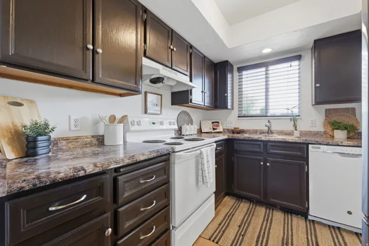 Kitchen featuring dark wood-finish cabinetry, white appliances, and a window with blinds above the sink