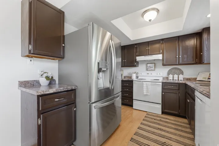 Stainless steel French door refrigerator, wood-finish flooring, dark wood-finish cabinetry, light-colored countertops, and a white electric range with overhead ventilation
