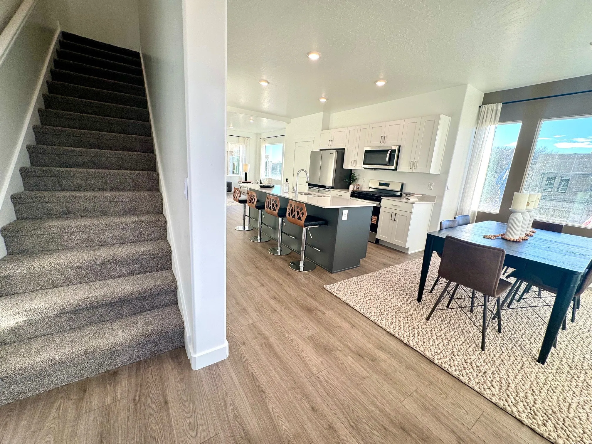 Open-concept living area featuring wood-finish flooring, a kitchen island with seating, white cabinetry, stainless steel appliances, and a dining area with expansive windows