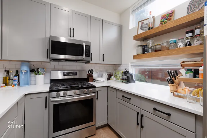 Gray shaker-style cabinetry with black hardware, stainless steel appliances, white subway tile backsplash, white countertops, and wood-finish open shelving