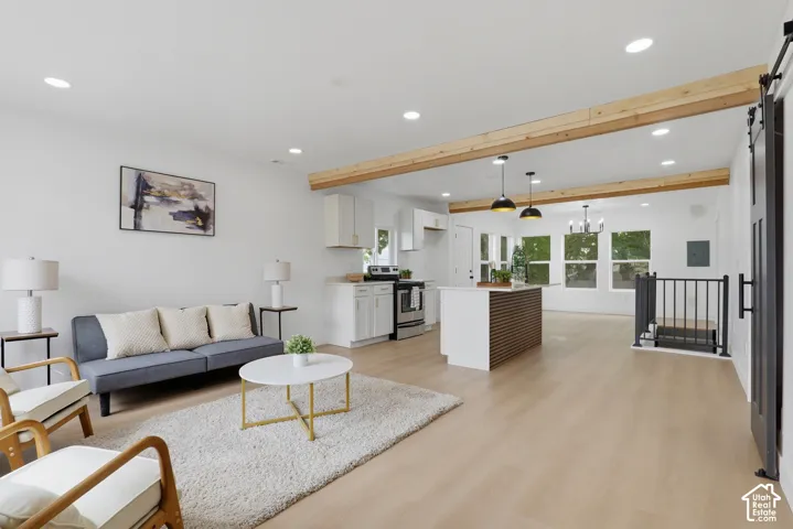 Open-concept living and kitchen space featuring exposed wood beam ceiling details, recessed lighting, light wood-finish flooring throughout, and large multi-pane windows