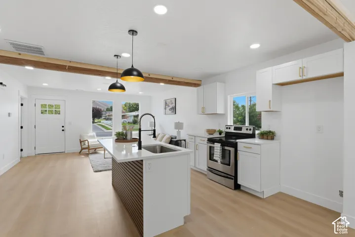 Open-concept living area featuring light wood-finish flooring, white cabinetry with gold-tone hardware, stainless steel appliances, and exposed wood beams