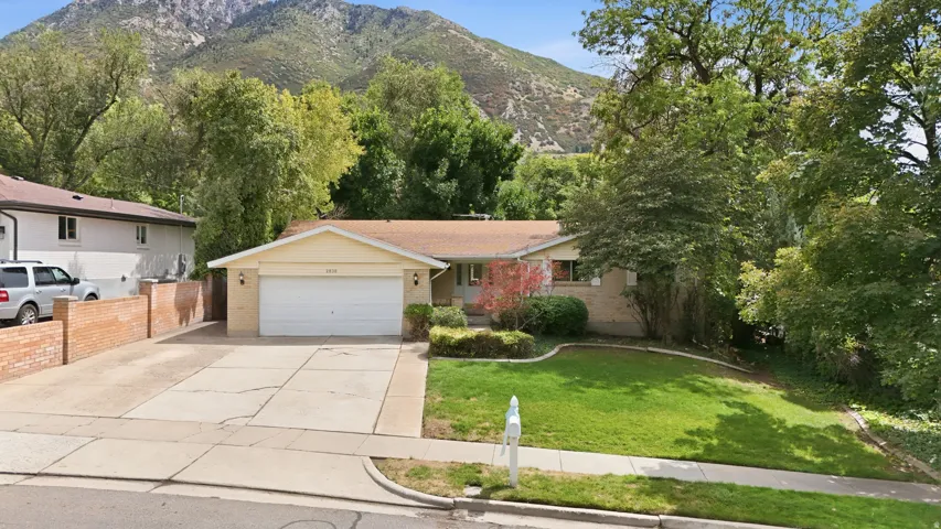 Ranch-style exterior with brick accents, an attached two-car garage, mature front yard landscaping, and a mountain backdrop
