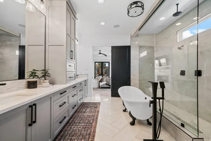 Dual vanities with integrated sinks, light gray cabinetry, a freestanding bathtub with dark-finish hardware, a glass-enclosed shower with a bench, and a chevron-pattern tile floor