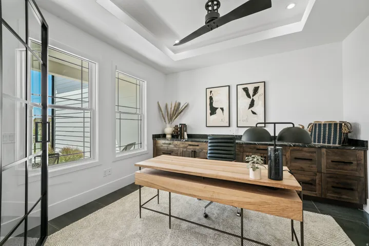 Recessed ceiling with a modern ceiling fan, built-in dark wood cabinetry with a dark countertop, steel-framed glass door, and two large windows