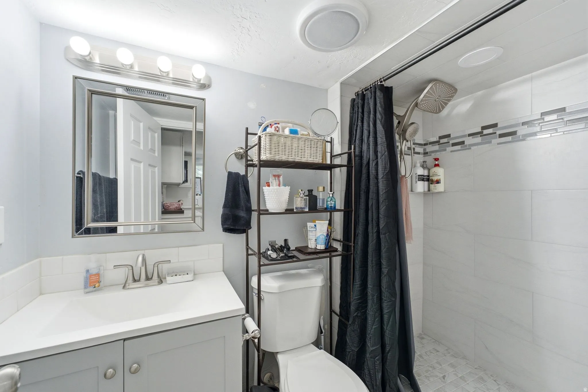 White subway tile backsplash, light gray vanity with brushed nickel hardware, framed mirror, white shower tiling with decorative accent band, and a multi-function showerhead