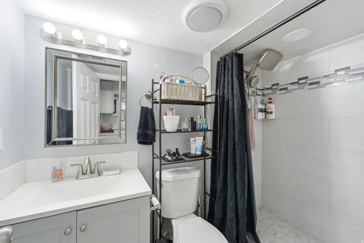 White subway tile backsplash, light gray vanity with brushed nickel hardware, framed mirror, white shower tiling with decorative accent band, and a multi-function showerhead