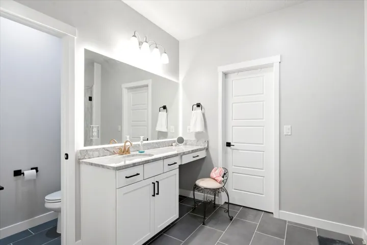 White vanity with a light-colored countertop, gold-finish faucet, and a large illuminated mirror