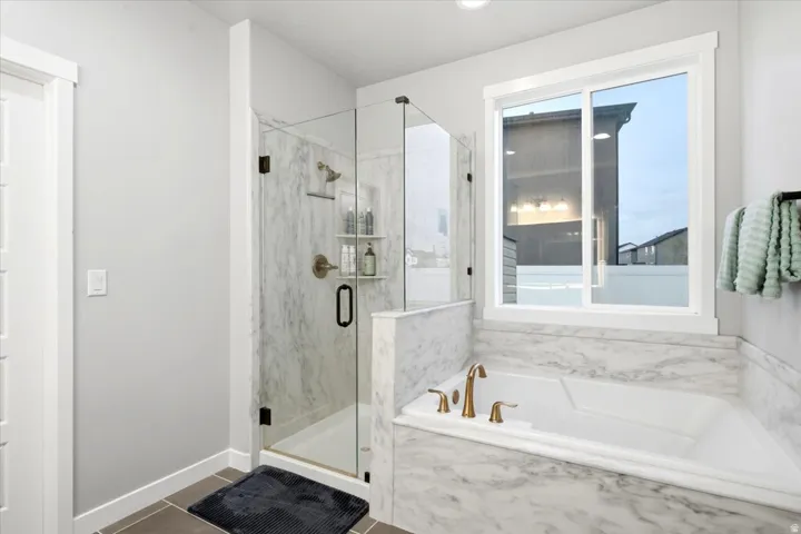 Walk-in shower with clear glass enclosure and built-in shelving, white soaking tub with gold-tone fixtures, marble-patterned tile surrounds, large window, and dark tile flooring