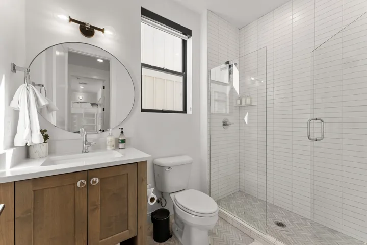 Bathroom featuring a wood-finish vanity with a white countertop, an oval mirror, and a glass-enclosed shower with a herringbone tile floor and white subway tile walls