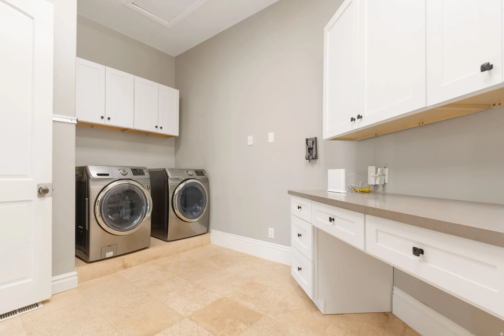 Light-toned tile flooring, white shaker cabinetry with dark hardware, integrated countertop workspace, and light gray wall paint