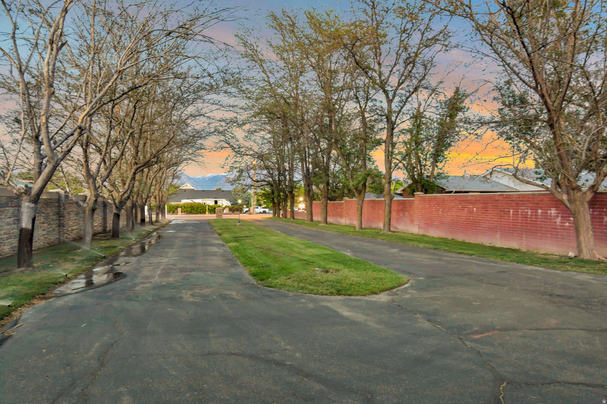 Paved drive with a central grassy median, lined by mature trees and a brick privacy wall