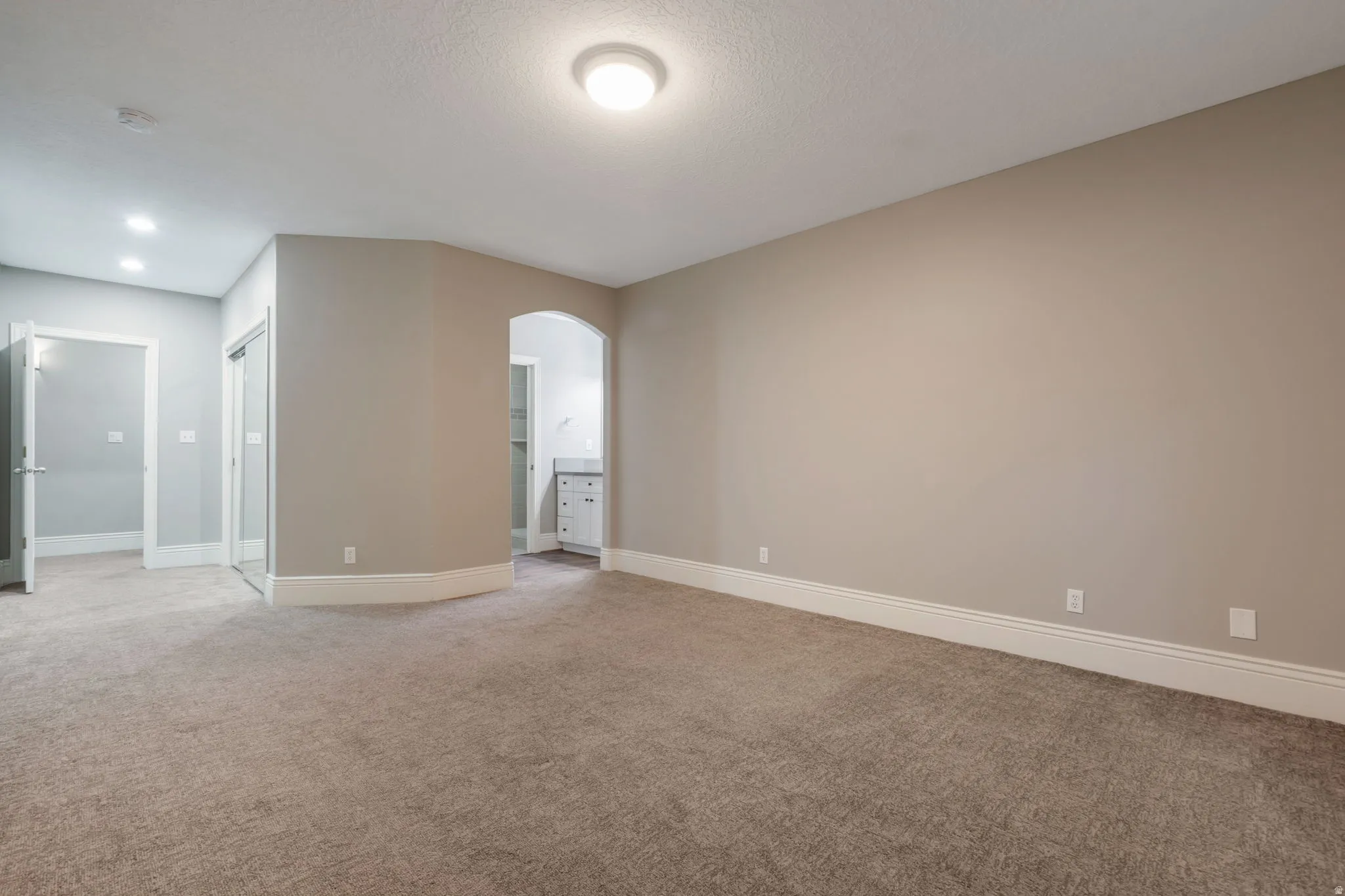 Carpeted room with neutral wall tones, recessed lighting, and an arched entry to an adjoining space featuring a vanity
