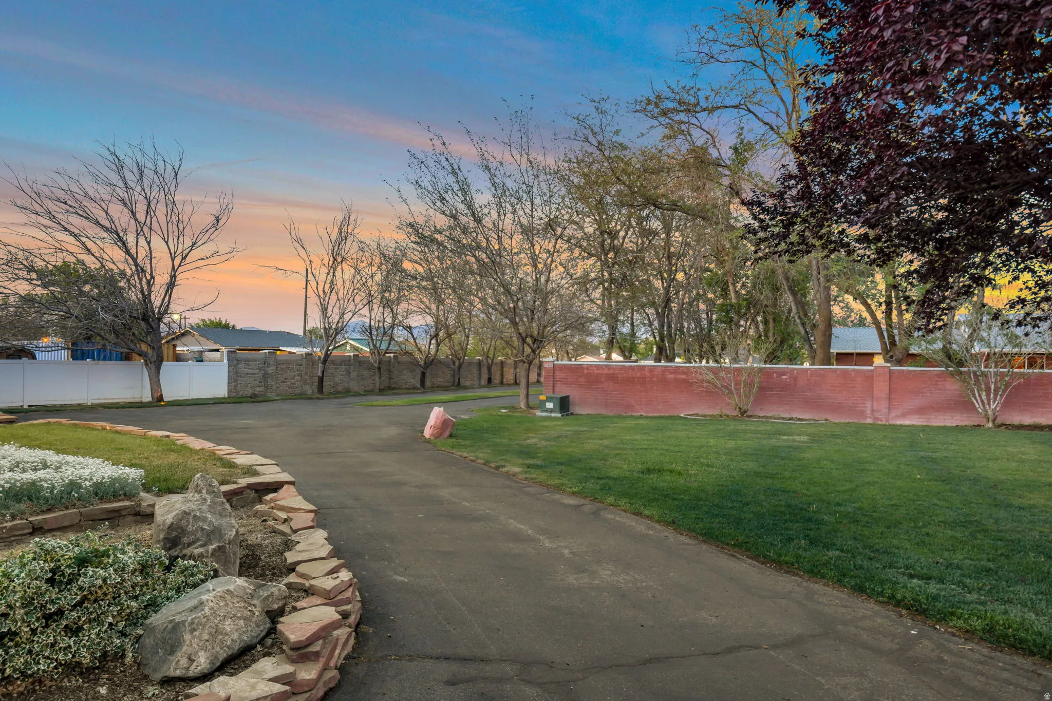Paved driveway bordered by a stone-edged planter with mature shrubs, leading to a landscaped lawn