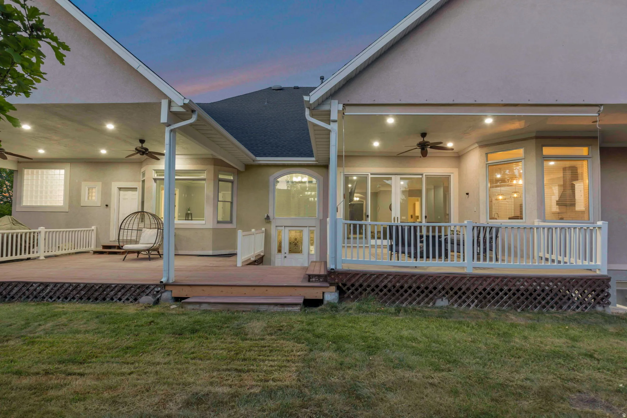 Expansive multi-level wood-finish deck with white railing, covered patio areas featuring recessed lighting and ceiling fans, and multiple large windows for abundant natural light