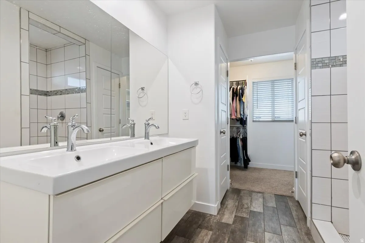 Dual basin vanity with chrome fixtures, large wall-mounted mirror, wood-finish flooring, and a tiled shower with decorative accent border