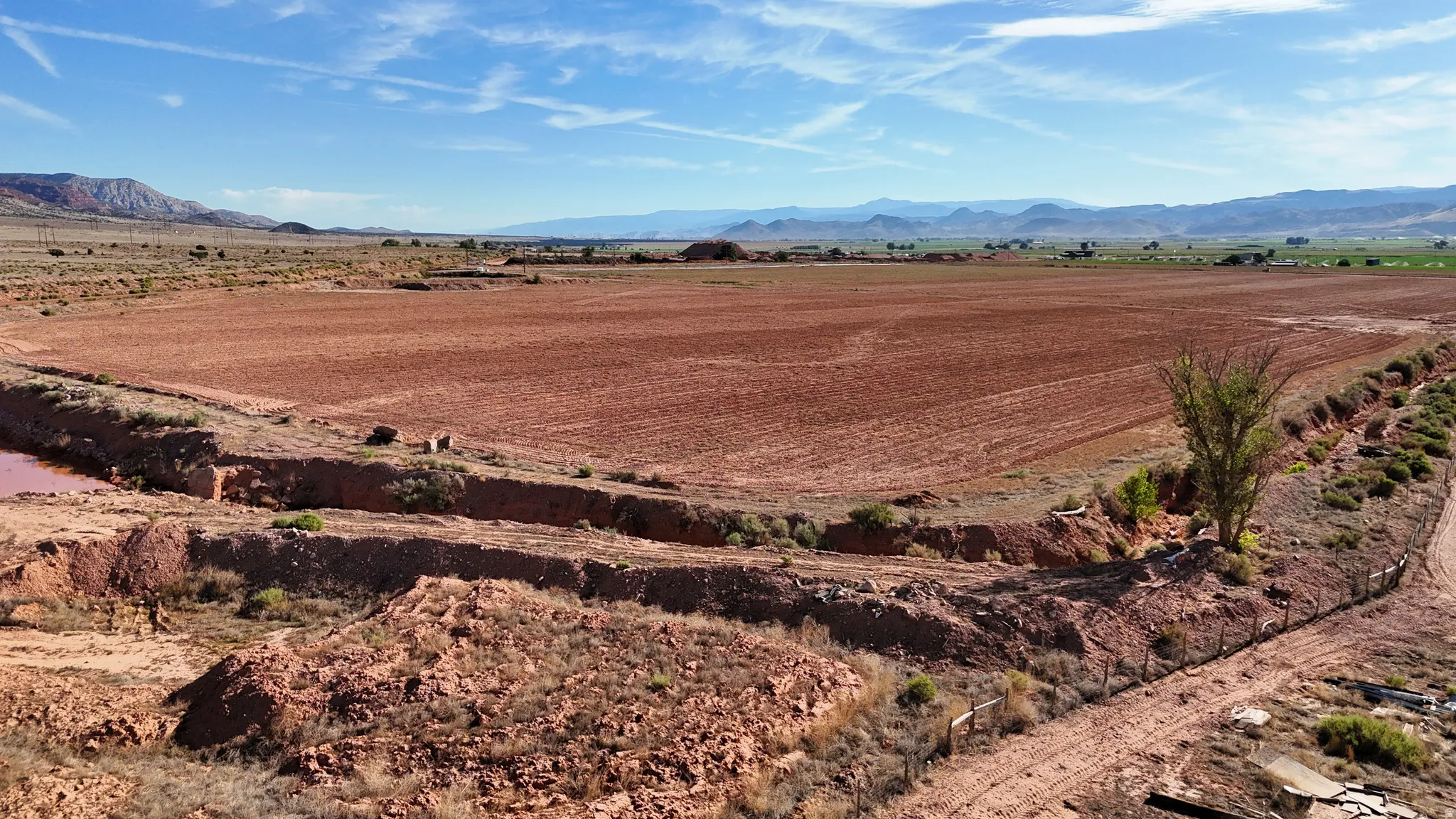Expansive tilled agricultural land with a natural water feature, bordered by mature vegetation and a dirt access road