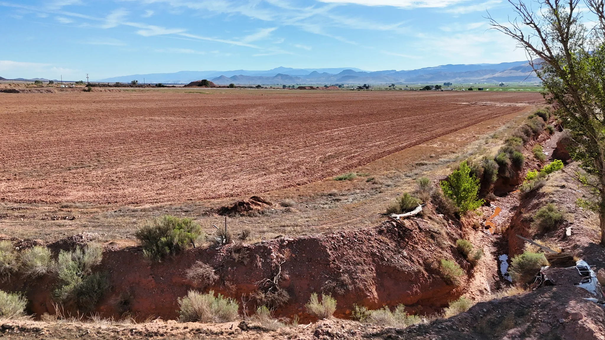 Expansive agricultural land featuring a natural water channel, desert vegetation, and distant mountains under a blue sky