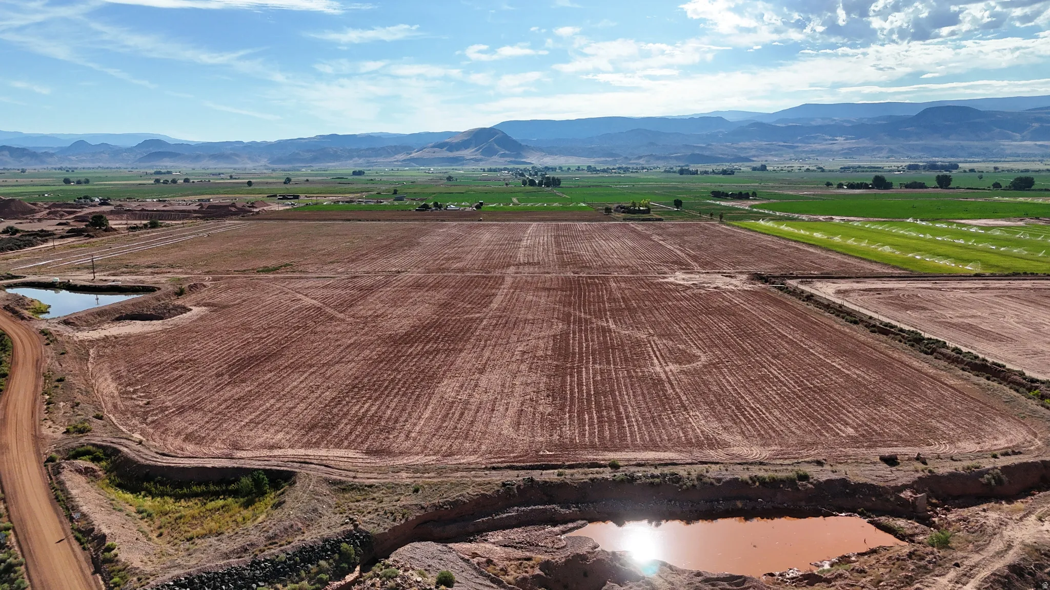 Expansive land featuring multiple tilled fields, two distinct ponds, a dirt road, and distant mountain ranges
