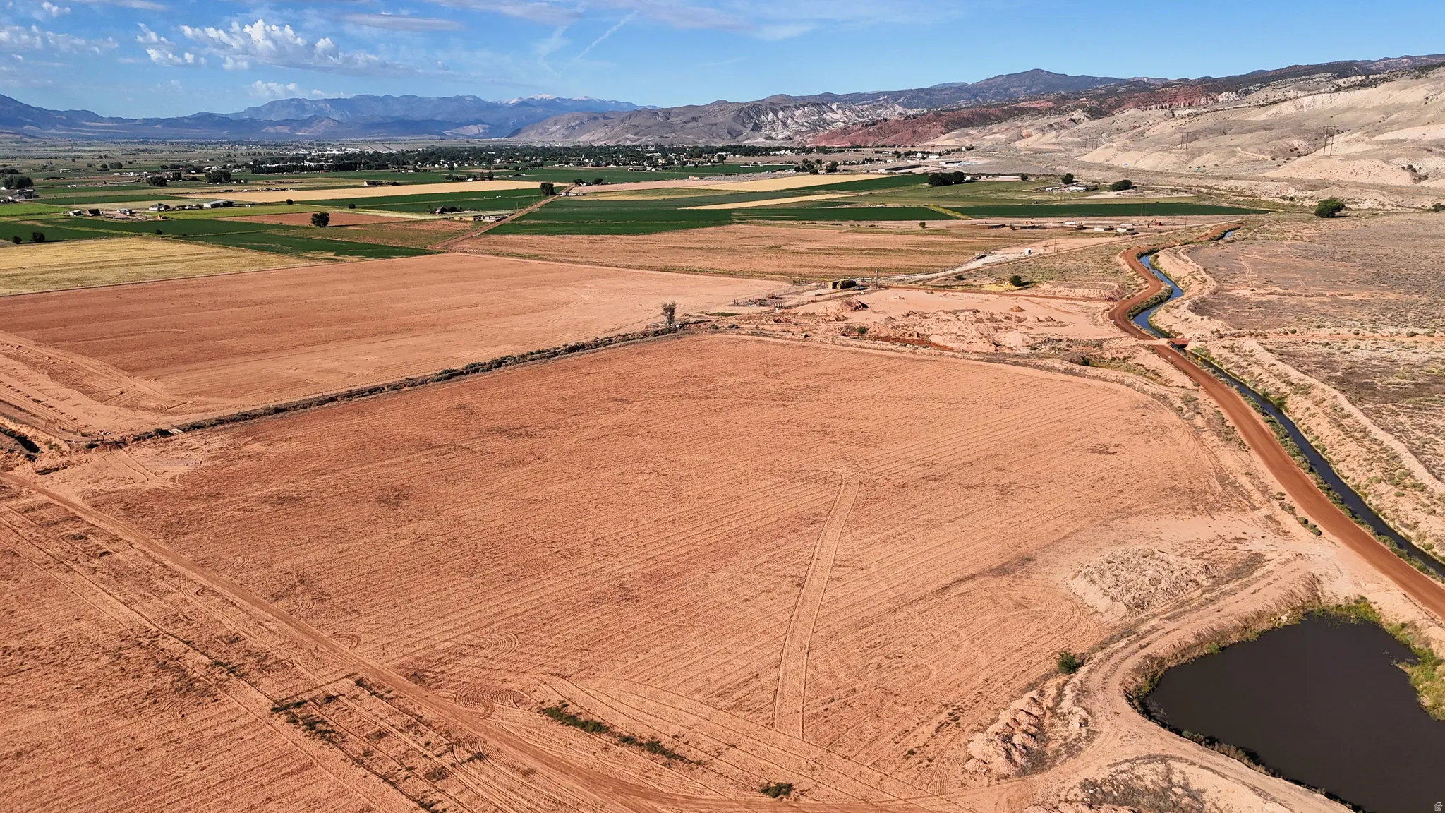 Expansive tilled land with visible irrigation channels, a water retention pond, and distant mountain backdrops