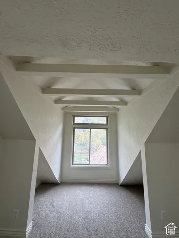 Vaulted ceiling with exposed decorative beams, wall-to-wall carpeting, and a large window featuring outdoor scenery