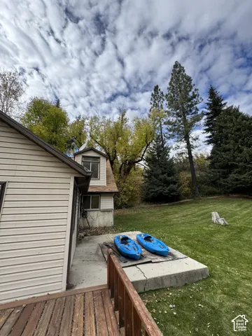 Exterior featuring horizontal siding, a raised wood-slat deck with railing, a concrete patio, and a grass-covered lot with mature trees