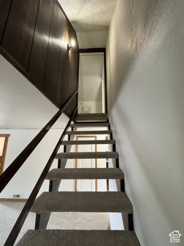 Staircase featuring neutral carpeted treads, metal handrails, dark wood-finish paneling, and a geometric-patterned ceiling