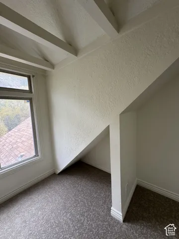 Vaulted ceilings with exposed beams, textured walls, neutral carpeting, and a multi-pane window