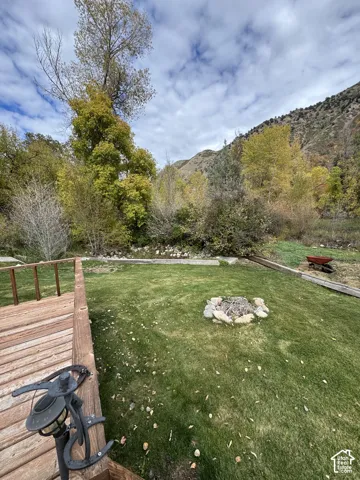 Backyard featuring a wood deck, stone-bordered fire pit, lawn, and mature trees against a mountain backdrop