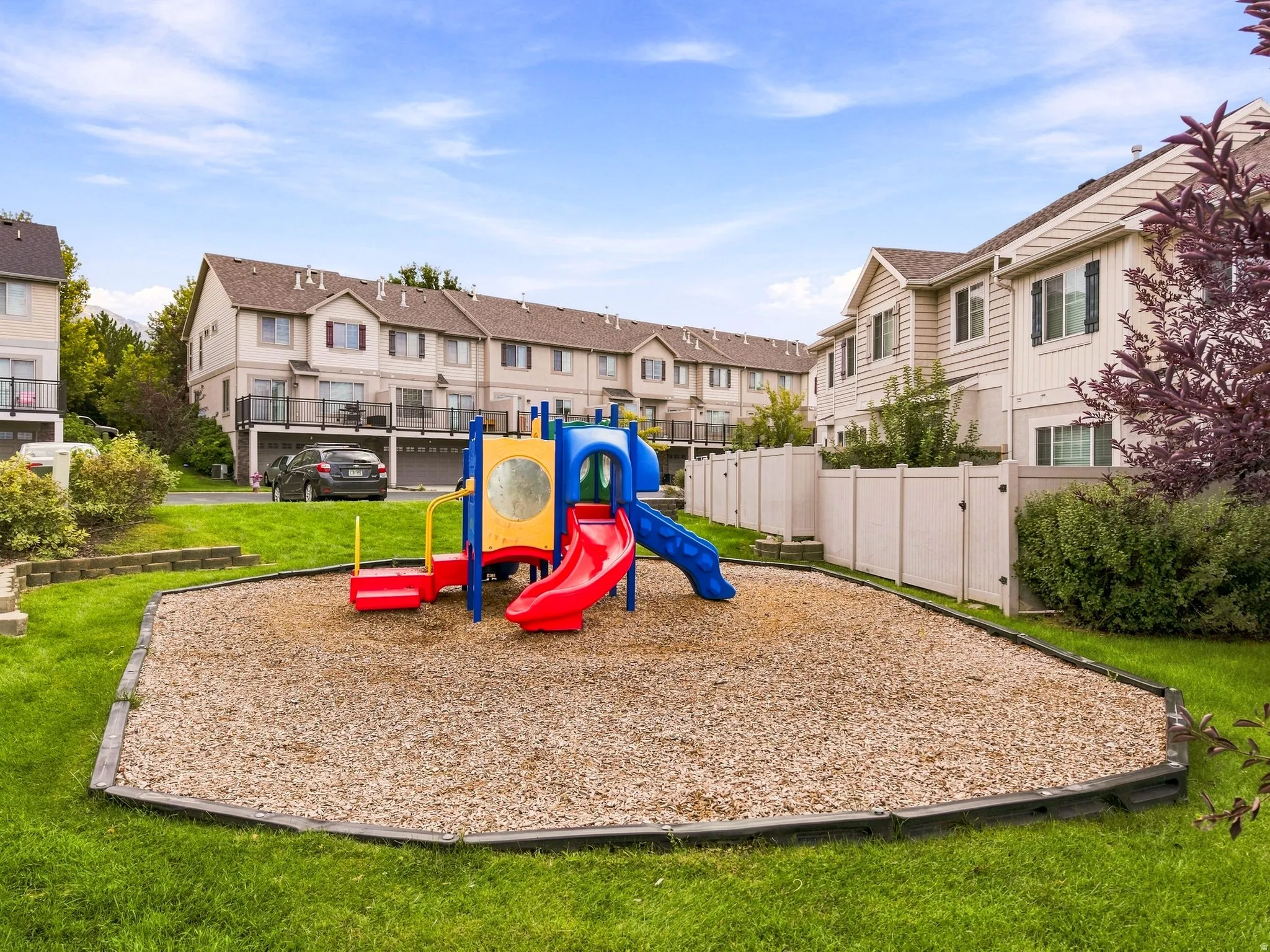 Community playground with wood chip ground cover, bordered by green lawn and a vinyl fence