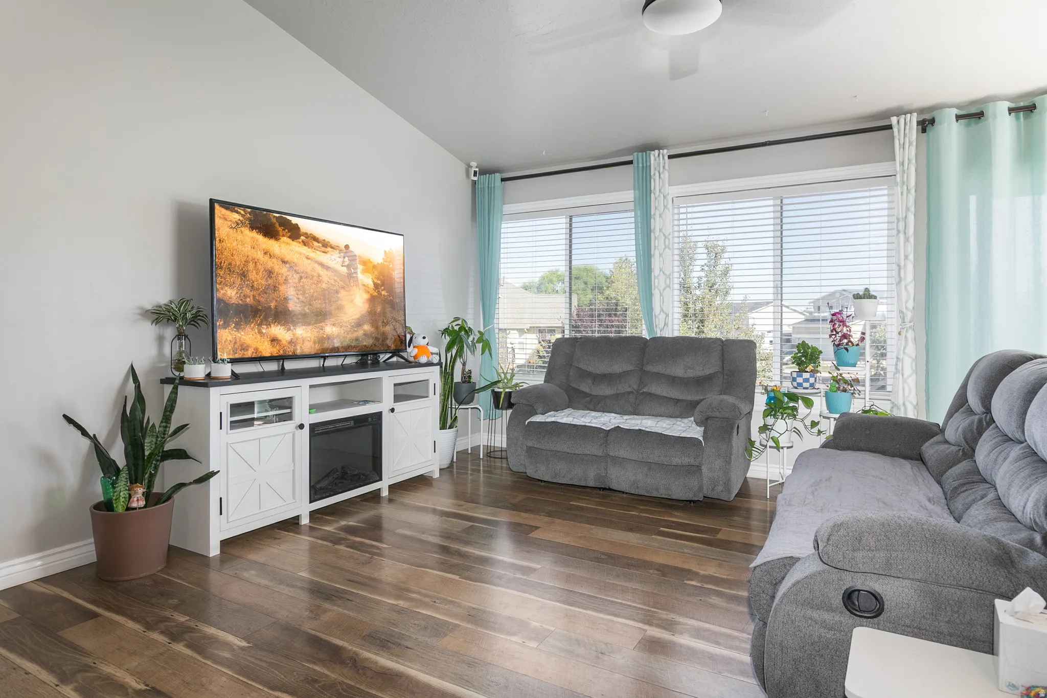 Vaulted ceiling, wood-finish flooring, white media console with integrated electric fireplace, and large windows with horizontal blinds