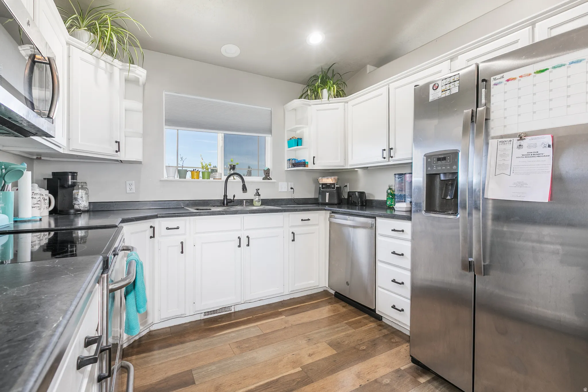 Kitchen featuring white cabinetry, dark countertops, wood-finish flooring, stainless steel appliances, and a window above the sink