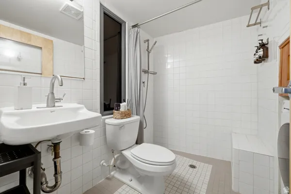 White tiled bathroom featuring a wall-mounted sink, toilet, and shower area with a built-in bench