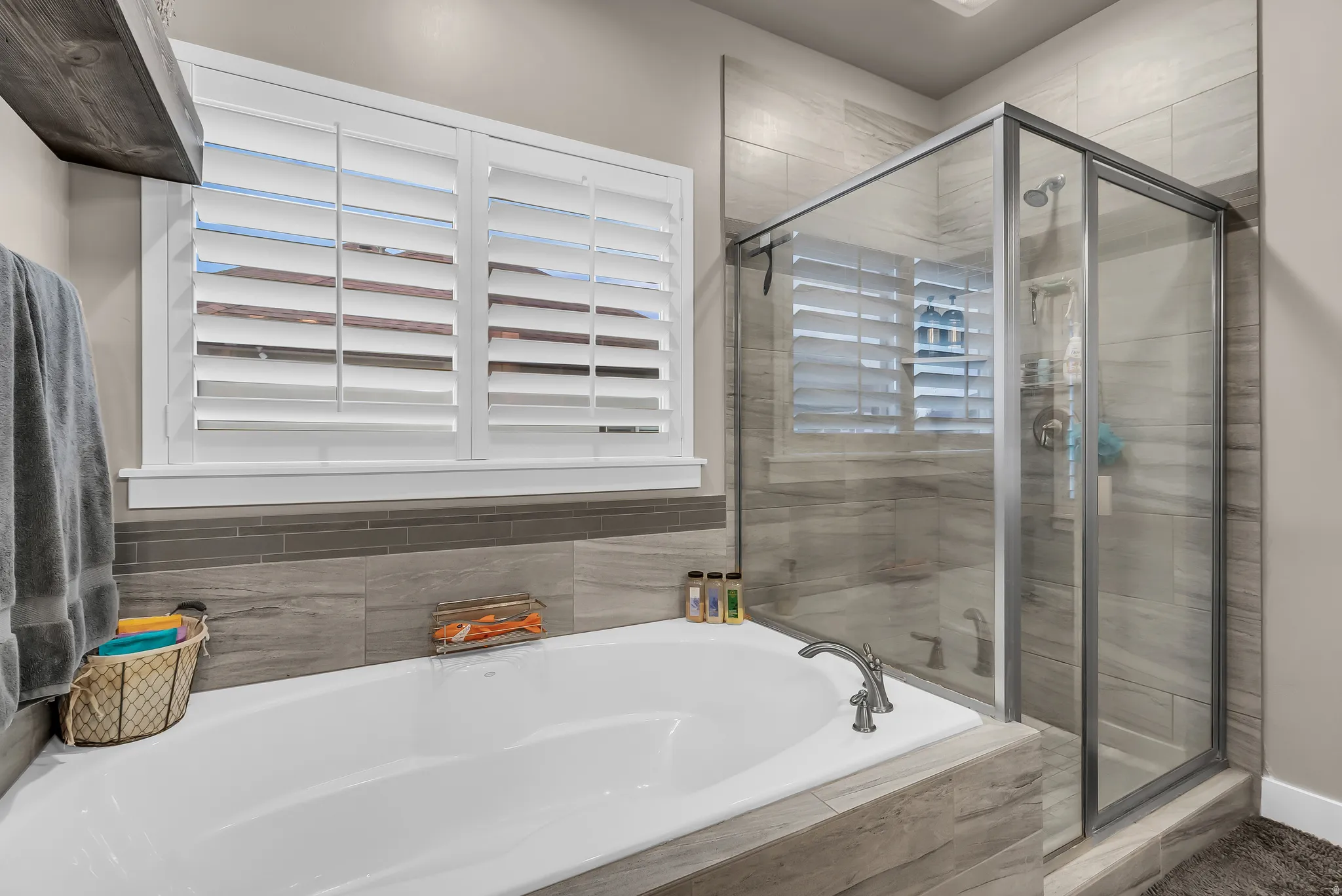 Built-in soaking tub, walk-in shower with glass enclosure, wood-finish tile surround, white plantation shutters, and brushed nickel fixtures