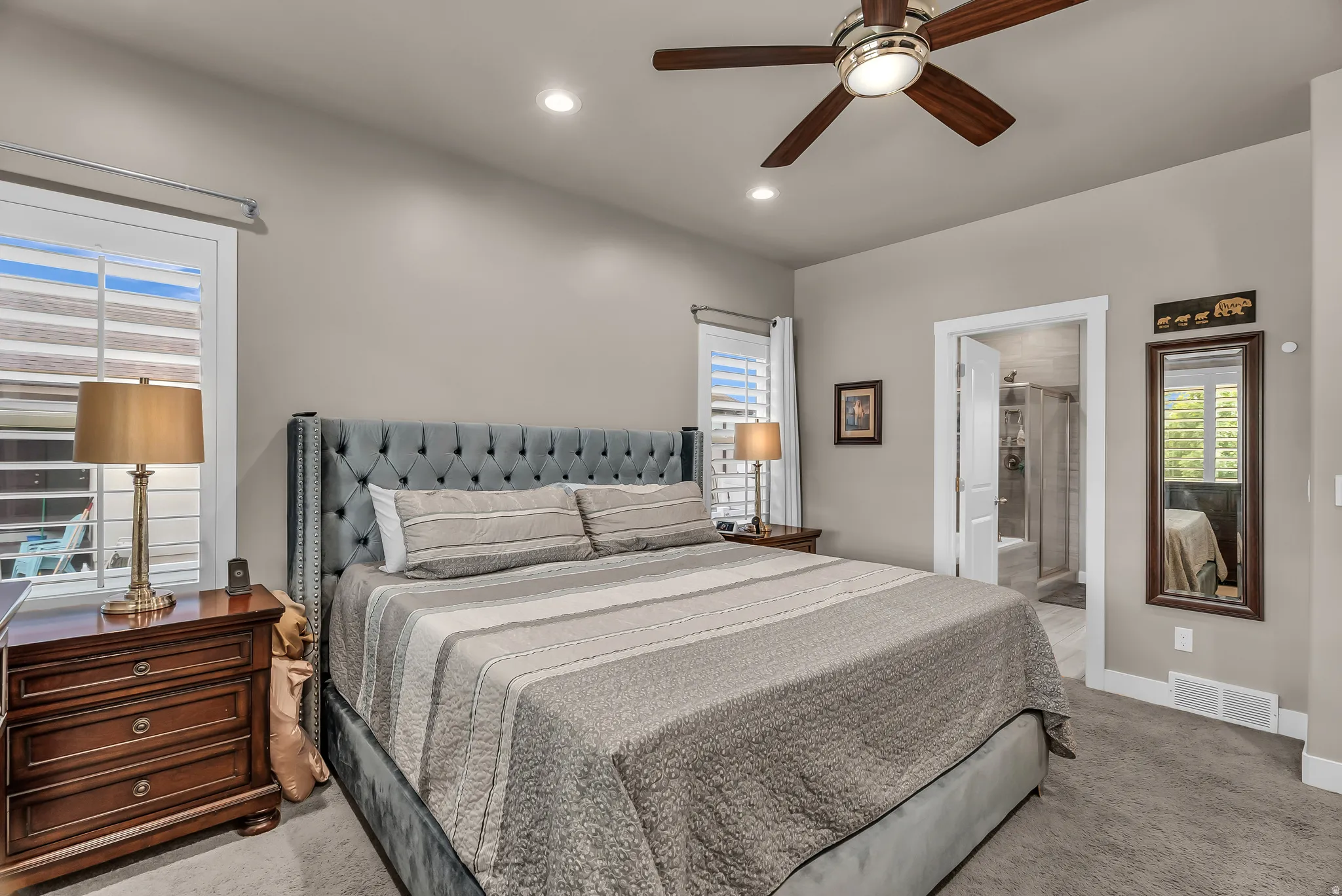 Recessed lighting, ceiling fan with light fixture, window with plantation shutters, carpeted flooring, and a doorway to a bathroom with tile flooring and a glass-enclosed shower