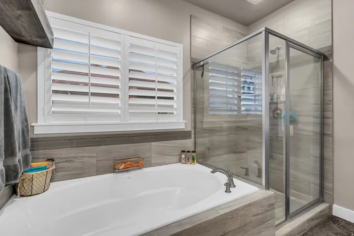 Built-in soaking tub, walk-in shower with glass enclosure, wood-finish tile surround, white plantation shutters, and brushed nickel fixtures