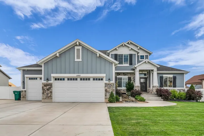 Two-story residence featuring a blue siding and stone facade, three garage doors, a covered entry with columns, and a landscaped front yard with a water feature