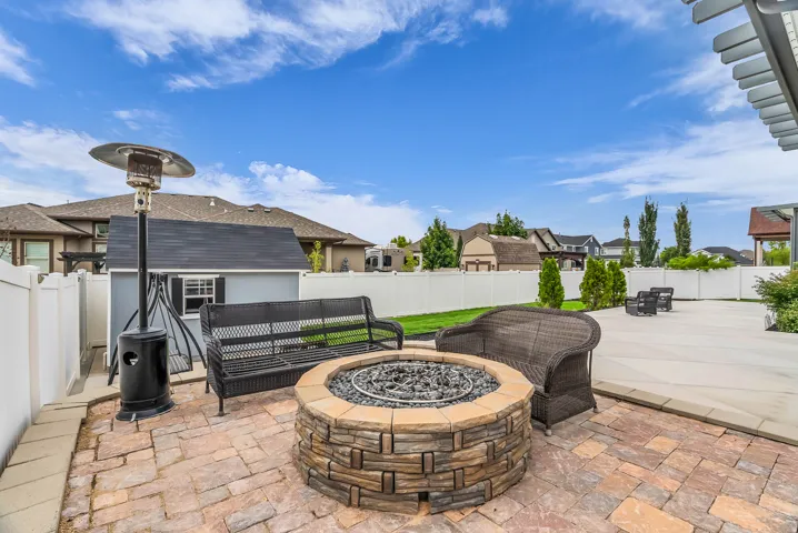 Paver patio with a built-in stone fire pit, extensive concrete patio, manicured lawn, and a white privacy fence