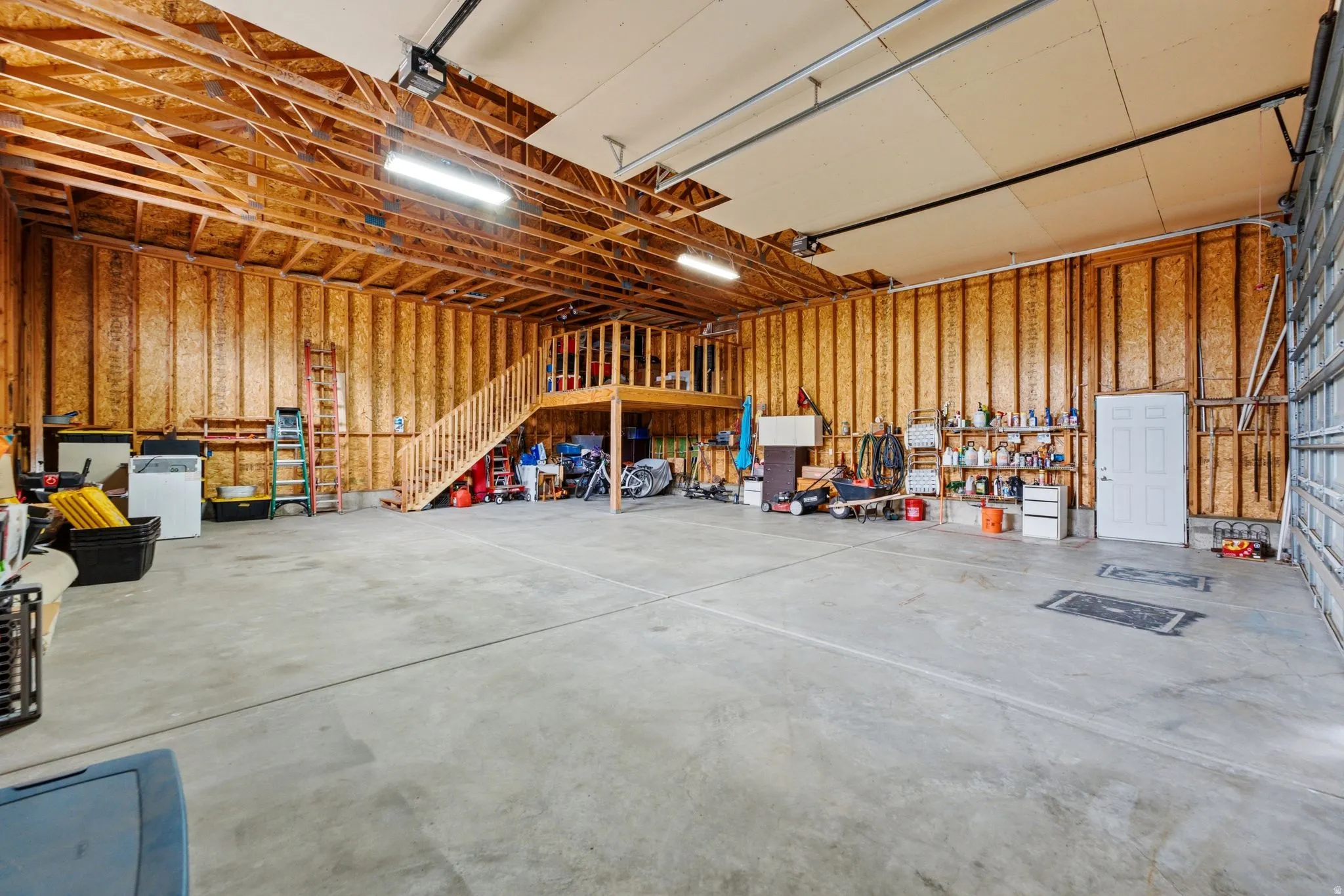 Spacious garage featuring a concrete floor, exposed wood framing, an interior staircase to a mezzanine, and a roll-up garage door