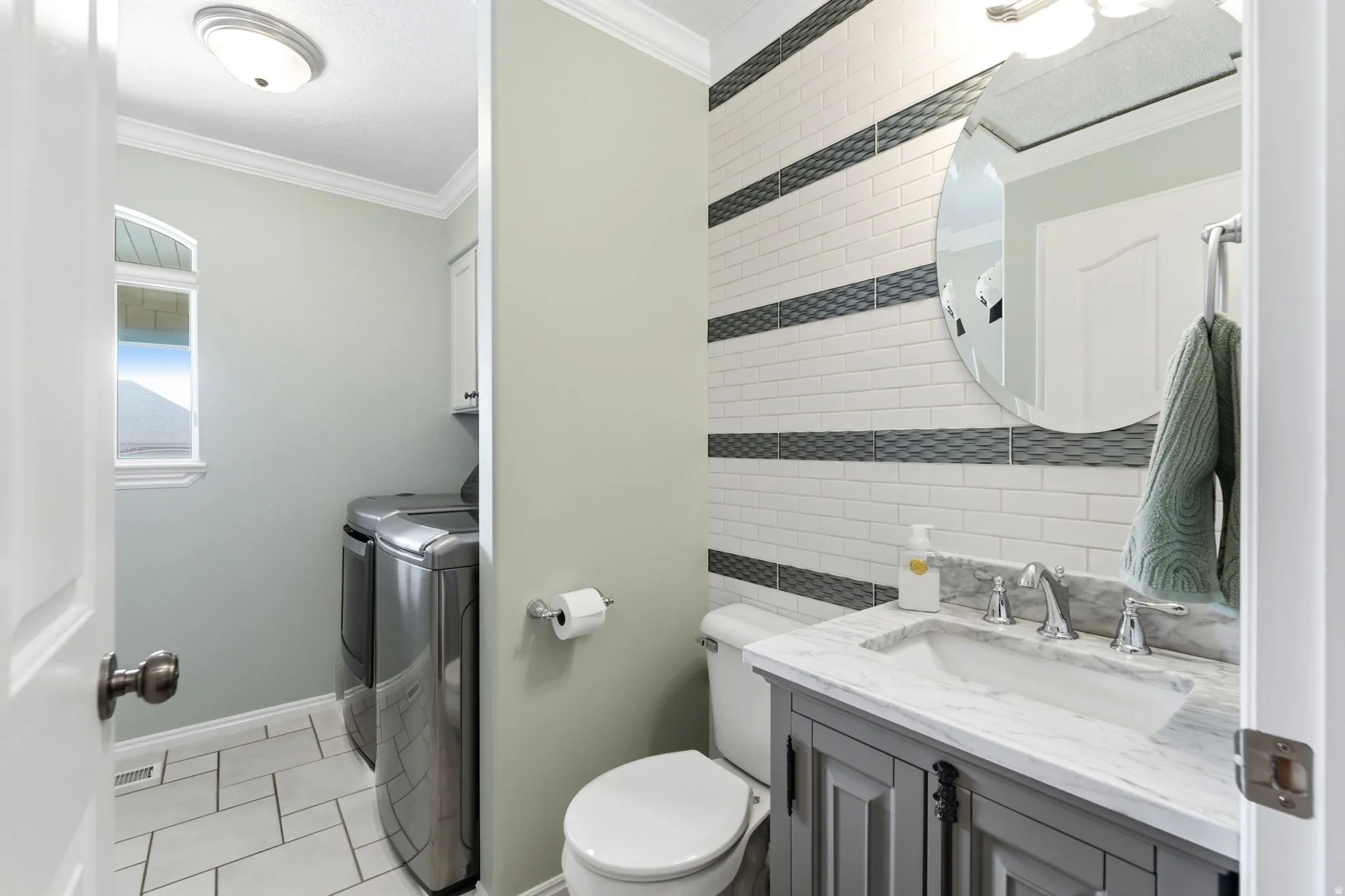 Vanity with integrated sink and chrome faucet, subway tile accent wall, circular mirror, crown molding, and tiled flooring