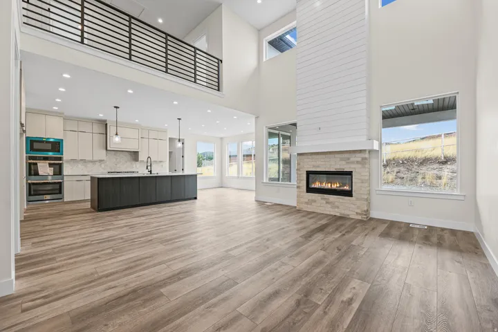 Double-height living space featuring wood-finish flooring, a contemporary fireplace with brick surround, an open-concept kitchen with a dark-toned island, and upper cabinetry with integrated appliances, all under a second-story interior balcony with metal