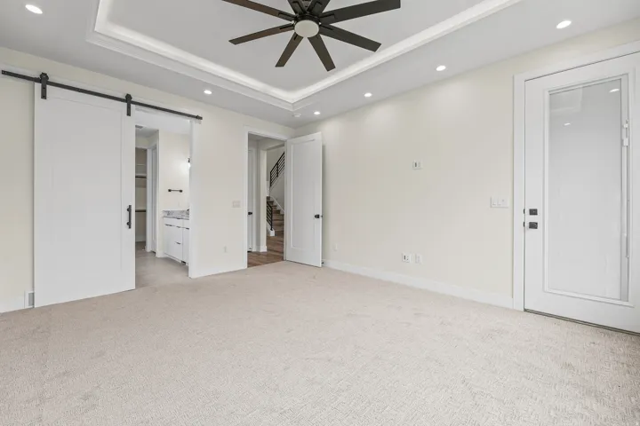 Tray ceiling with recessed lighting and a ceiling fan, light-toned carpeting, white barn door, white paneled door with glass insert, and white solid paneled door