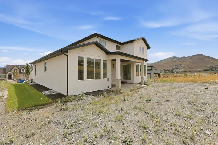 Contemporary stucco exterior featuring a covered patio, multiple large windows, stone-clad columns, and a grassy side yard