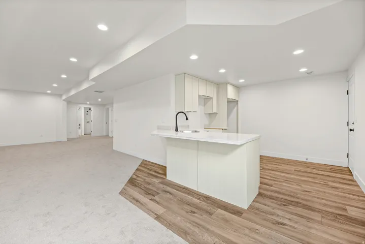 Spacious room featuring recessed lighting, a built-in kitchenette with light-toned cabinetry, a kitchen island with an undermount sink and matte black faucet, and a combination of carpet and wood-finish flooring